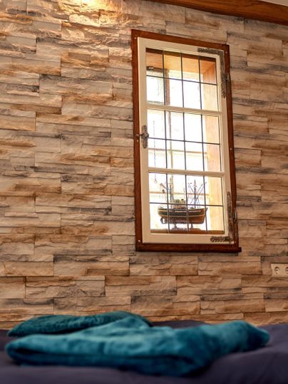 A room with a stone wall and a window. On the bed lies a blue cloth.