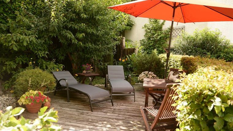 An inviting garden with two loungers and a red parasol. Surrounded by lush greenery and flowers, the space invites relaxation.