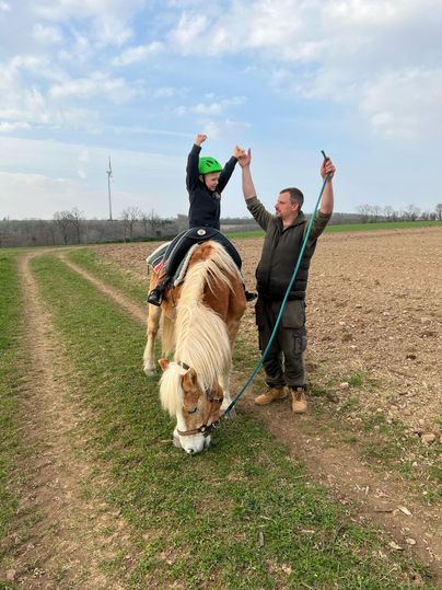 A child is riding a horse and raising their arms in celebration. An adult is holding the horse by the reins and smiling.