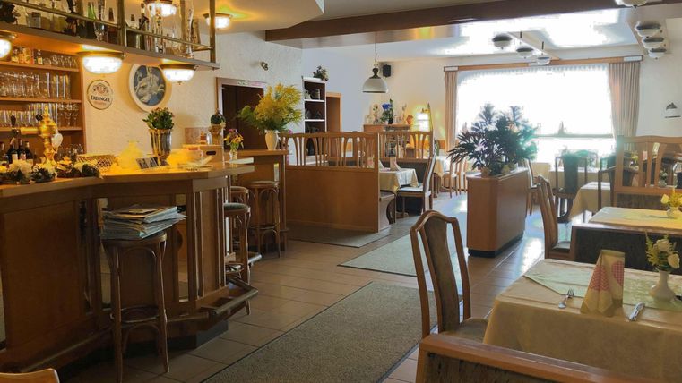 Ein einladendes Restaurant mit Holzmöbeln und gemütlicher Atmosphäre. Die Fenster bieten Blick auf eine grüne Pflanze und Tageslicht strömt herein.