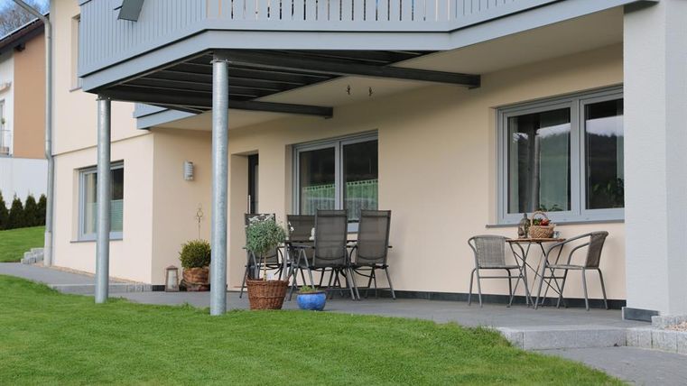 Eine moderne Terrasse mit Gartenmöbeln und einem kleinen Tisch. Der Rasen ist gepflegt und die Wand des Hauses ist in einem hellen Farbton gestrichen.