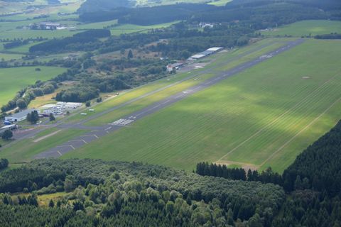 Een uitgestrekt vliegveld met groen gras en een blauwe lucht. Op de achtergrond zijn bomen en enkele gebouwen zichtbaar.