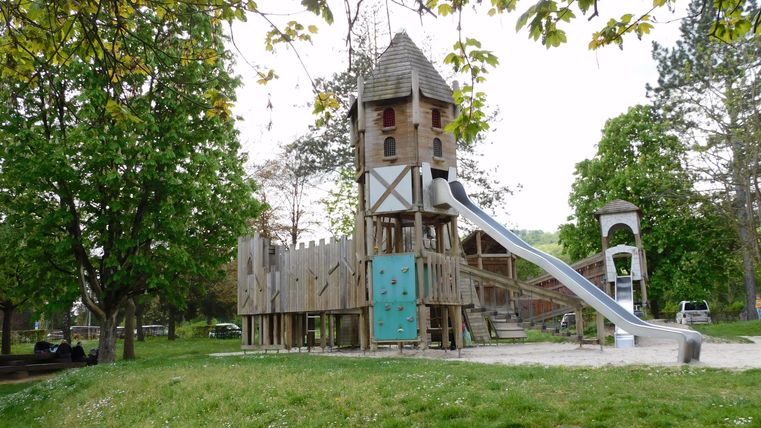 Ein Spielplatz mit einem großen Holzspielturm und einer Rutsche. Umgeben von Bäumen und Grünflächen.