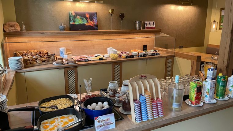 Ein reichhaltiges Frühstücksbuffet mit Eiern, Speck und verschiedenen Brötchen. An der Theke stehen auch Getränke und Süßigkeiten bereit.