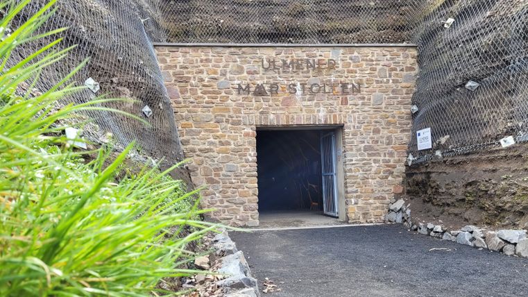 Ein Eingang zu einer Steinmine, umgeben von einer steinernen Wand und Netzschutz. Grüne Pflanzen wachsen in der Nähe des Eingangs.