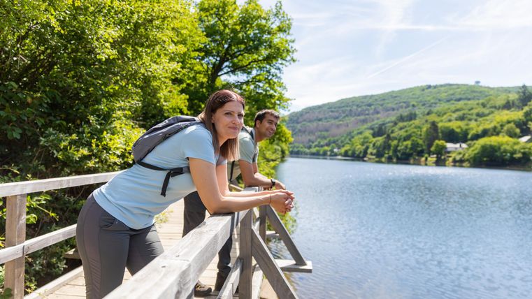 Zwei Personen stehen an einem Steg und genießen die Aussicht auf einen ruhigen Fluss. Umgeben von üppigem Grün und sanften Hügeln strahlt die Szenerie Ruhe und Naturverbundenheit aus.