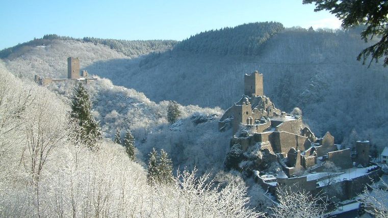 Eine winterliche Landschaft mit schneebedeckten Bäumen und Hügeln. Im Vordergrund sind Ruinen einer alten Burg sichtbar.