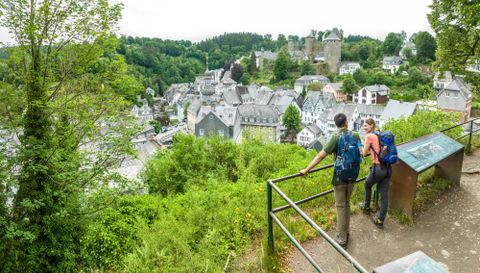 Eine malerische Aussicht auf ein kleines Dorf mit historischen Gebäuden und einer Burg im Hintergrund. Zwei Wanderer genießen die Aussicht von einem Aussichtspunkt umgeben von grüner Natur.