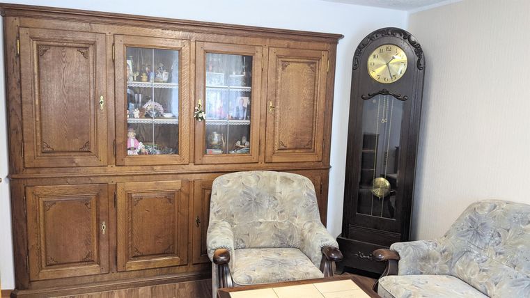 A cozy living room with a wooden cabinet and two chairs. In the background, there is a large wall clock.