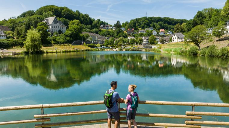 Ein Paar steht an einem Ufer und blickt auf einen ruhigen See. Im Hintergrund sind grüne Hügel und Häuser zu sehen.