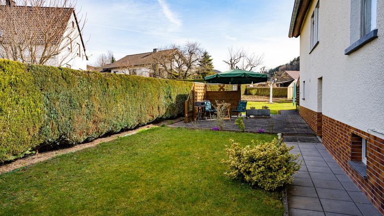 Ein gepflegter Garten mit einer grünen Hecke und einer Terrasse. Im Hintergrund steht ein Sonnenschirm und einige Gartenmöbel sind sichtbar.