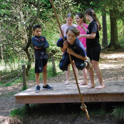 Een groep kinderen speelt in het bos. Een jongen zwaait aan een touw, terwijl de anderen kijken.