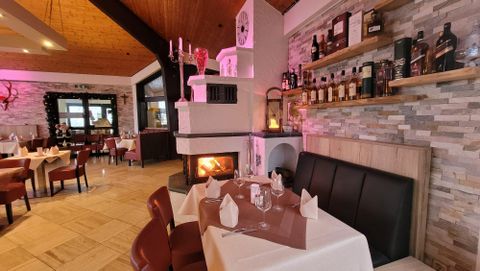 A cozy restaurant with wooden tables, a fireplace, decorative bottle shelves, and set tables. The atmosphere is warm and inviting.