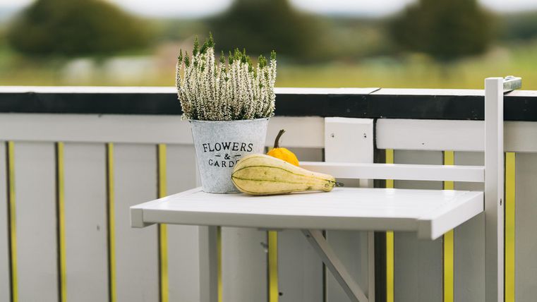 Een kleine tafel op een balkon met een bloemenplant in een bloempot en een gele banaan. Op de achtergrond zijn vage bomen te zien.