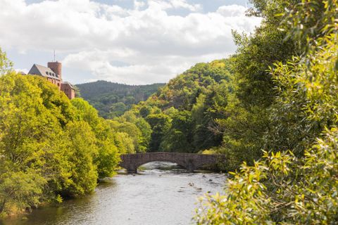 Een schilderachtig landschap met een rivier en een boogbrug. Omringd door groene bomen en zachte heuvels onder een bewolkte lucht.