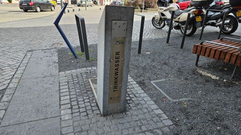 Een drinkwatertap van beton staat op een plein. Op de achtergrond zijn motorfietsen en zitbanken zichtbaar.