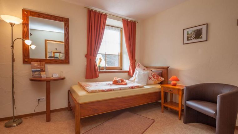 Ein gemütliches Zimmer mit einem Einzelbett und einem Stuhl. Die Wände sind in hellen Farben gehalten und die Fenster sind mit roten Vorhängen dekoriert.