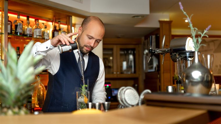 Ein Barkeeper mixt einen Cocktail an einer eleganten Bar. Im Hintergrund sind zahlreiche Spirituosen sichtbar.