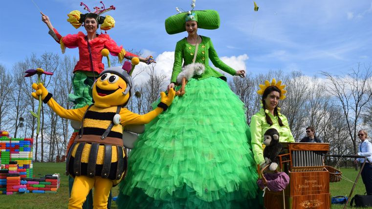 Eine fröhliche Gruppe von Straßenkünstlern in bunten Kostümen steht im Freien. Im Hintergrund ist ein blauer Himmel mit wenigen Wolken zu sehen.