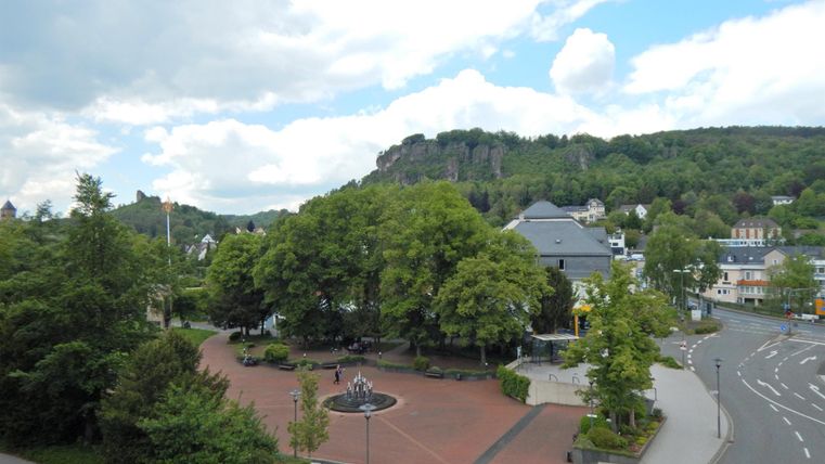 A quiet place with green trees and a central fountain. In the background, hills and a blue-and-white sky are visible.