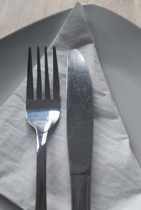 A place setting with a fork and a knife on a white napkin. In the background, a gray plate is visible.