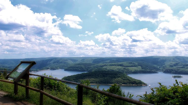 Eine atemberaubende Aussicht auf einen Fluss, umgeben von Hügeln und dichter Vegetation. Der Himmel ist blau mit vielen Wolken.