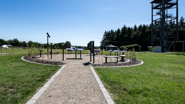 A well-maintained outdoor area with various informational stations and a viewing tower in the background. The landscape is green and sunny.