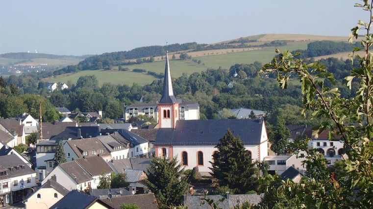 Blick von oben auf den Ort Stadtkyll, in deren Mitte die Kirche St. Josef steht.Das Gebäude ist im klassizistischen Stil errichtet,mit einem einfachen viereckigen Mittelschiff. Rechts ist der Glockenturm mit einem hohen, spitzen,schwarz eingedecktem Dach. Das Gebäude ist in weiß gestrichen, die Rundfenster sind, genauso wir die Ecken des Glockenturm ,rötlich abgesetzt.