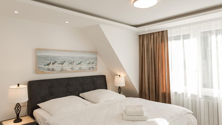 Ein gemütliches Schlafzimmer mit einem großen Doppelbett und zwei Nachttischen. Helle Farben und Fenster mit Vorhängen sorgen für eine entspannte Atmosphäre.