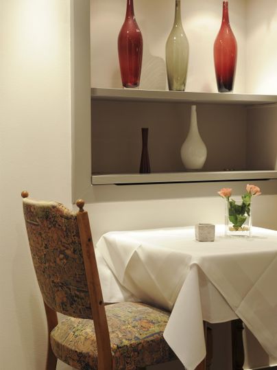 Ein eleganter Tisch mit einer weißen Tischdecke und einer kleinen Blumenvase. Im Hintergrund sind dekorative Flaschen in verschiedenen Farben auf einem Regal platziert.