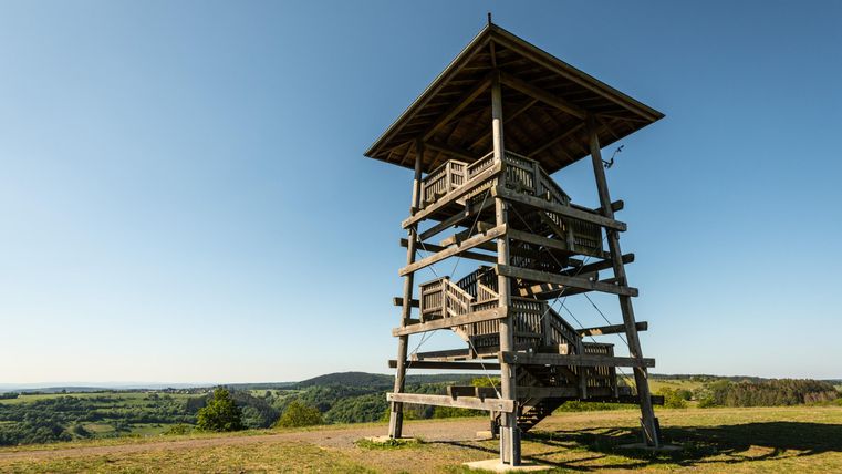 Ein Aussichtsturm aus Holz steht auf einem Hügel. Im Hintergrund sind sanfte Hügel und klarer Himmel zu sehen.