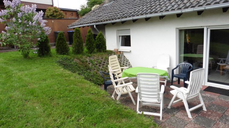 Ein gemütlicher Außenbereich mit einem Tisch und Stühlen auf einer grünen Wiese. Im Hintergrund ist ein Haus und ein blühender Strauch sichtbar.