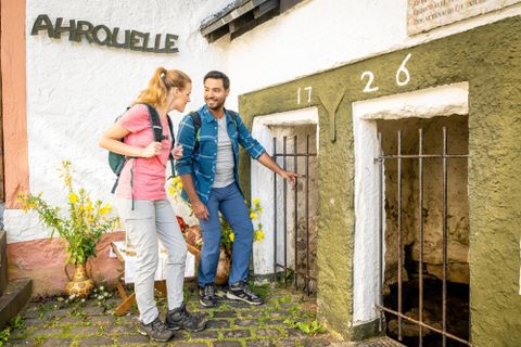 Zwei Wanderer stehen vor einem alten Brunnen mit zwei Eingängen, die mit Gittertüren versehen sind. Im Hintergrund ist ein Schild mit der Aufschrift "AHROUELLE" zu sehen.
