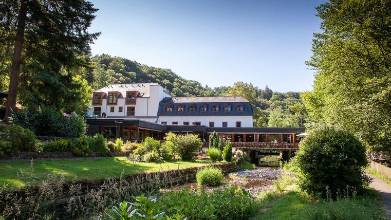 Ein schönes Hotel liegt am Rande eines ruhigen Gartens mit einem kleinen Teich. Umgeben von Bäumen und Hügeln bietet es eine entspannte Atmosphäre.