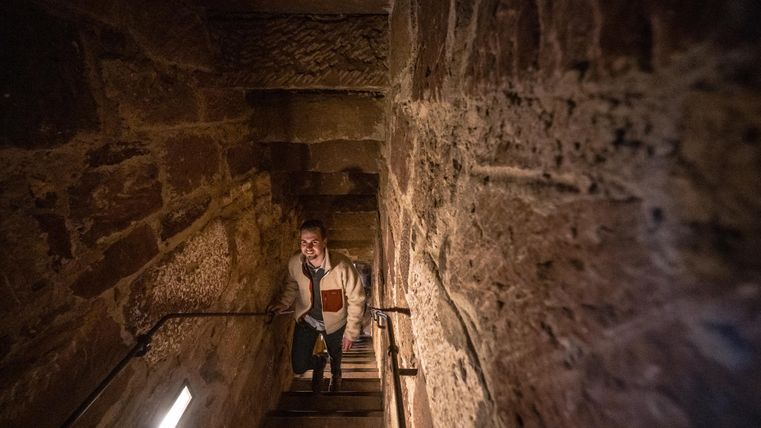 A person is climbing a stone staircase. The walls are made of rough stone and the light is dim.