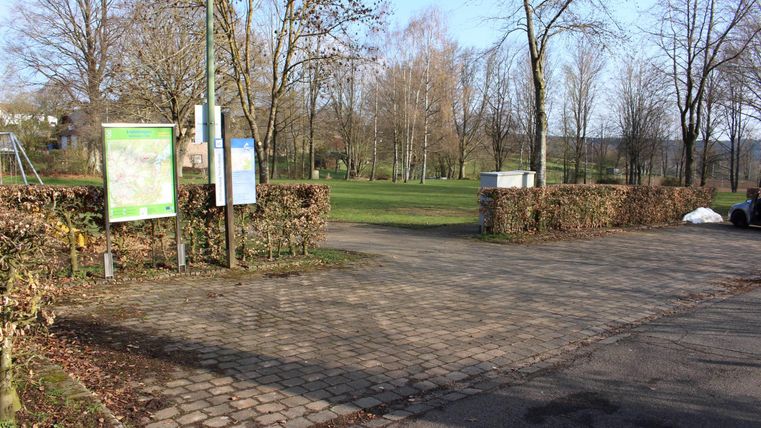 Een parkeerplaats die aan een park grenst, met banken en borden. Op de achtergrond zijn bomen en een heldere lucht te zien.