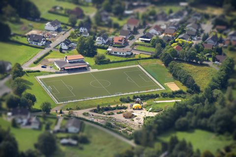 Ein Fußballplatz umgeben von Wohngebieten und grünen Wiesen. Im Vordergrund ist ein Spielplatz zu sehen.