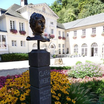 Eine Bronze-Büste von Clara Wieck in einem schönen Garten mit bunten Blumen. Im Hintergrund ist ein historisches Gebäude zu sehen.