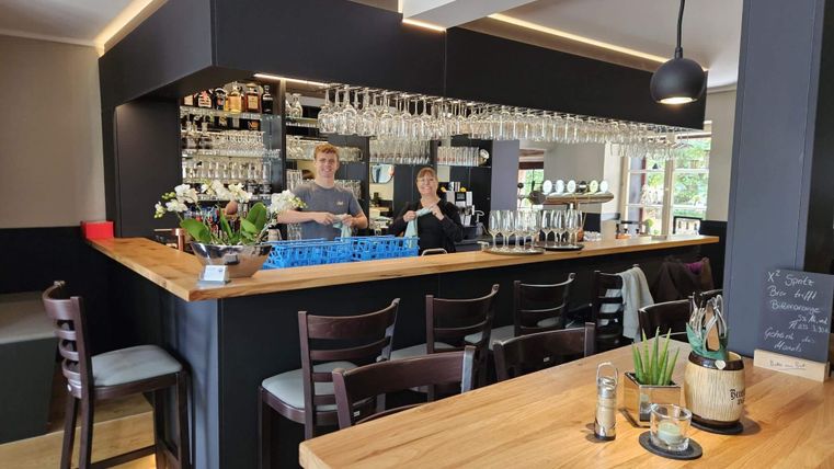 Een moderne bar met een houten toog en talloze glazen. Twee medewerkers staan vriendelijk achter de bar.