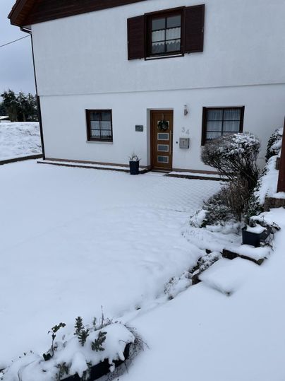 Een huis in de sneeuw met een besneeuwde oprit. De tuin is bedekt met sneeuw en er staan enkele planten in potten.