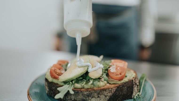 Ein gesunder Toast mit frischem Gemüse und einer cremigen Sauce. Jemand führt gerade die Sauce über die Zutaten.