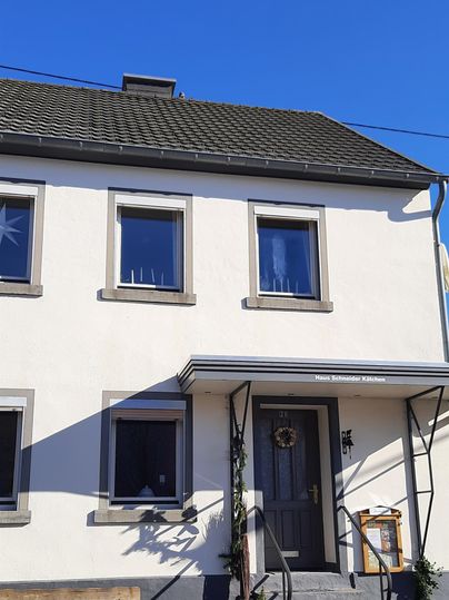 Ein modernes Haus mit einem schlichten weißen Fassadenanstrich und einem grauen Dach. Der Himmel ist klar und blau, und vor dem Haus befindet sich eine kleine Terrasse.
