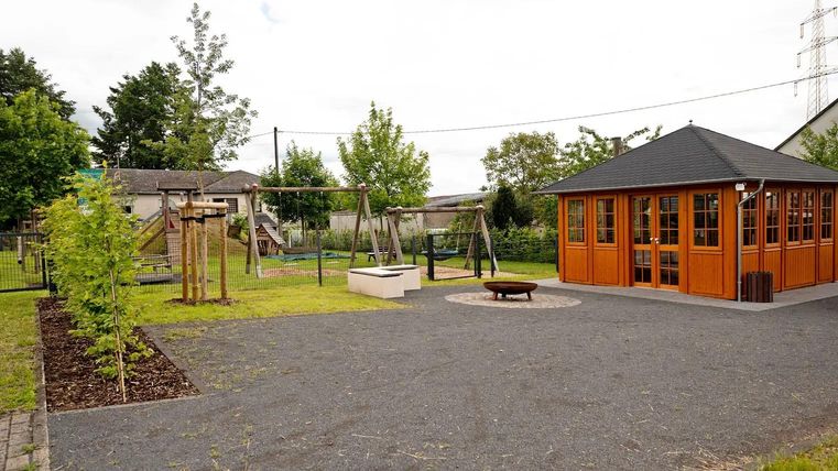 Een speelplaats met schommels en een houten huis. De groene omgeving is ingericht met bomen en een verhard oppervlak.