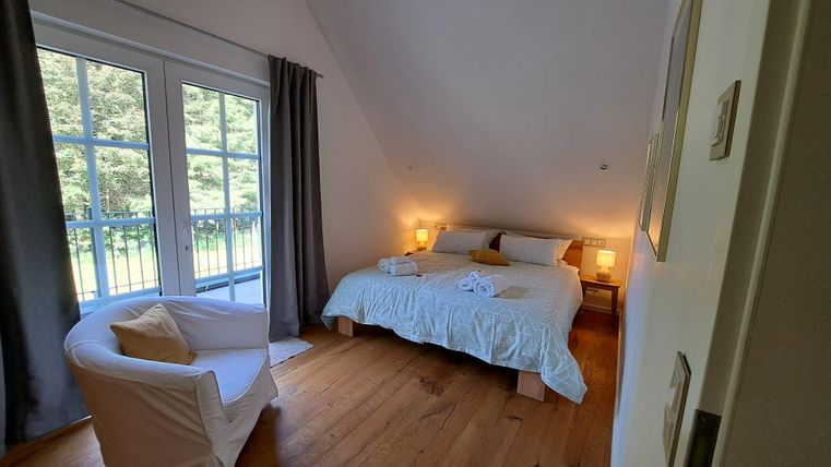 A cozy bedroom with a large bed and a comfortable armchair. Large windows provide plenty of daylight and a view of the greenery.