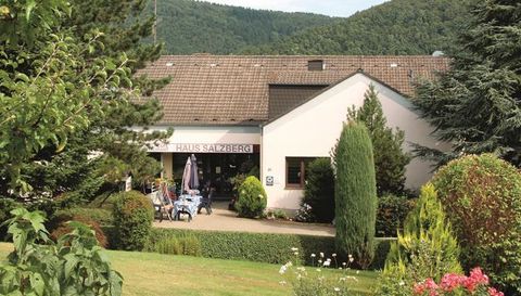 Ein Gebäude mit der Aufschrift "Haus Saltzberg" umgeben von Bäumen und bunten Blumen. Im Hintergrund sind sanfte Hügel sichtbar.