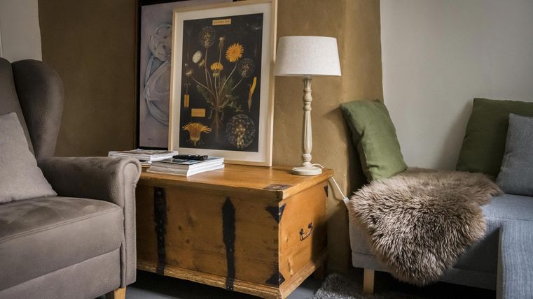 Ein gemütlicher Wohnraum mit einem bequemen Sessel und einem Sofa. Neben einem Holzschrank steht eine Lampe und ein Kunstwerk an der Wand.