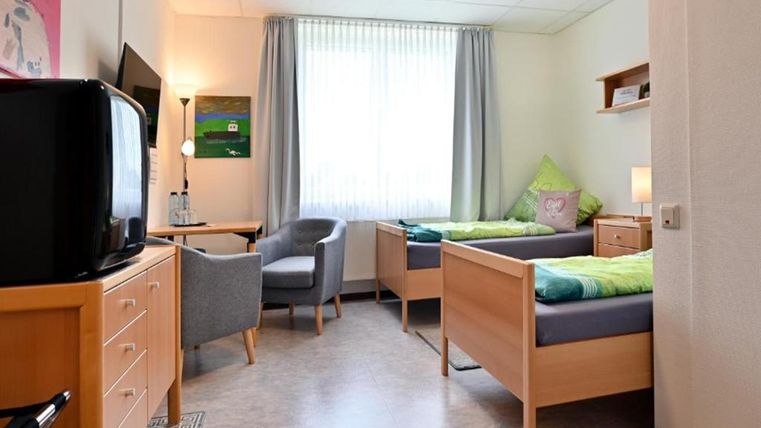 A simple room with two single beds and a window that lets in plenty of natural light. There is an armchair, a television, and a wardrobe for personal belongings.