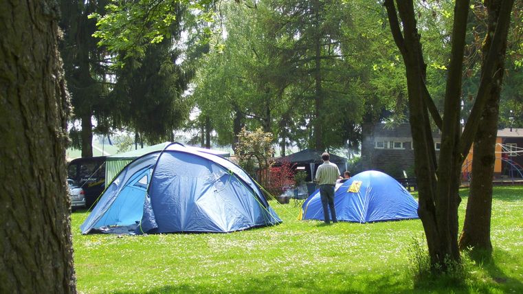 Een camping met blauwe tenten op groen gras. Omgeven door bomen en een zonnige lucht.