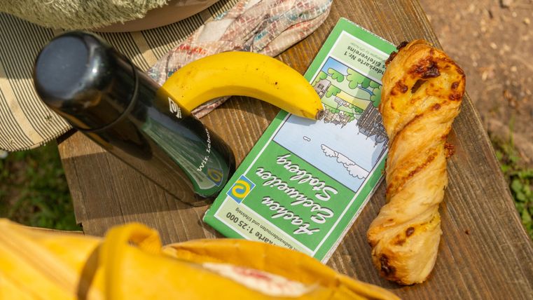 A picnic with a bread roll, a banana, and a thermos on a wooden table. The surroundings seem sunny and relaxing.