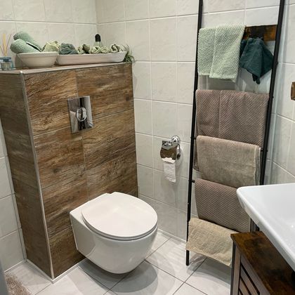 Een moderne badkamer met een witte toiletpot en een houten plank. Handdoeken in zachte kleuren hangen aan een standaard naast een wastafel.